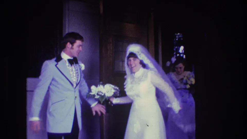 In 1974 Boston home-movie footage, a groom in a pale blue tuxedo and a bride in a white gown and veil stand at a building entrance with a bridesmaid partly visible behind them.