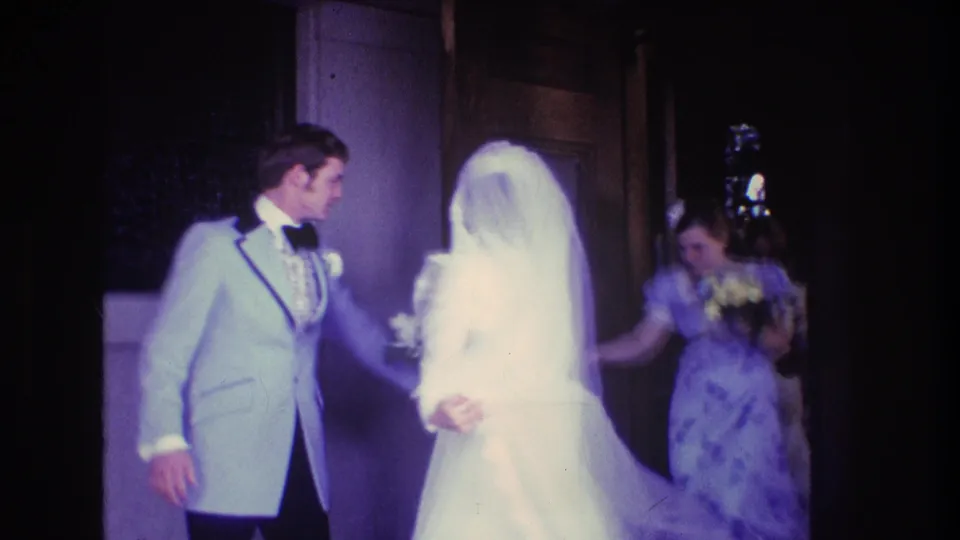 The groom turns toward the veiled bride at the doorway while a bridesmaid in a purple dress follows close behind in the 1974 Boston wedding footage.