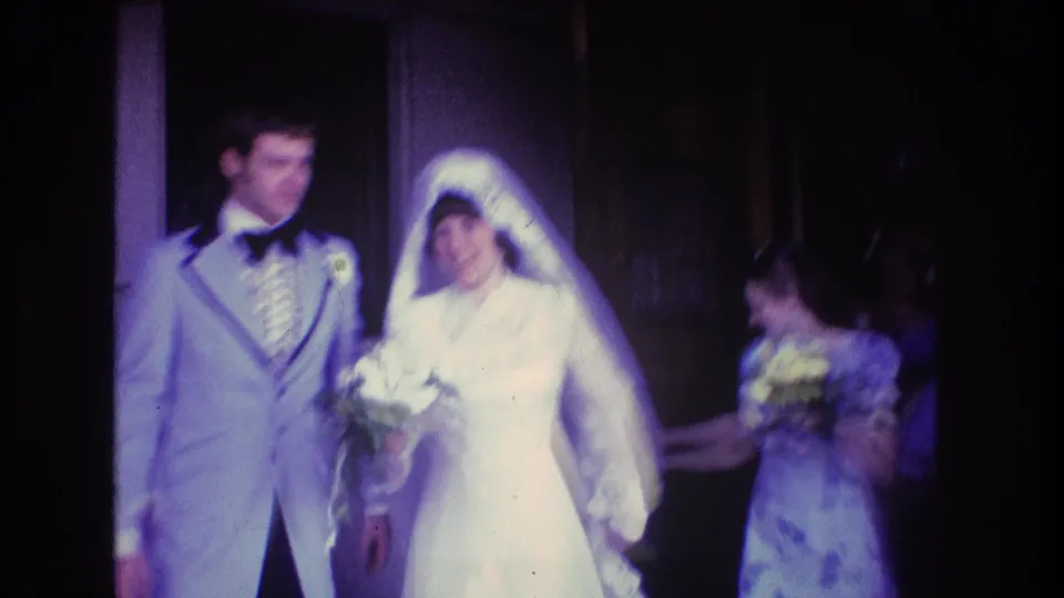 The bride and groom move forward from the entrance, with the bride holding a bouquet and a purple-dressed attendant visible at the side.