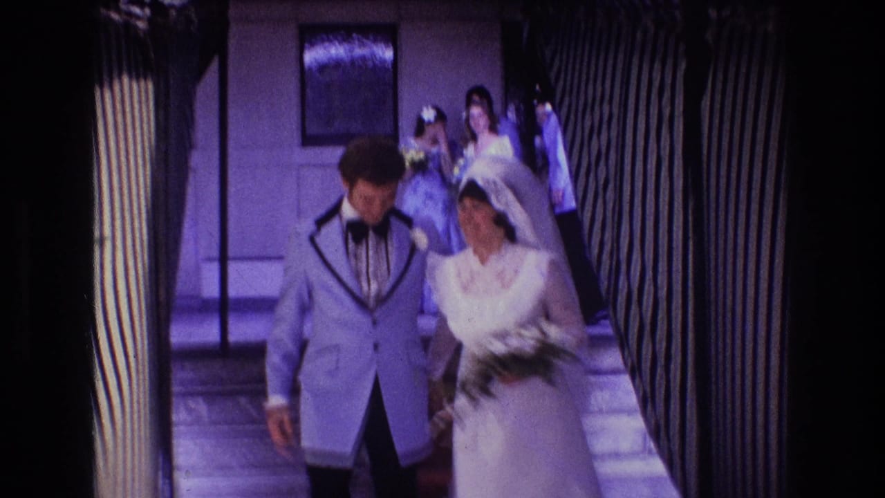 In 1974 Boston 8mm wedding footage, a bride in a white gown and veil walks beside a groom in a light blue tuxedo through a narrow striped hallway with guests behind them.