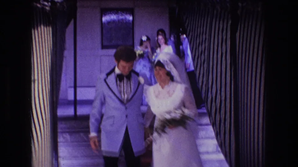 In 1974 Boston 8mm wedding footage, a bride in a white gown and veil walks beside a groom in a light blue tuxedo through a narrow striped hallway with guests behind them.