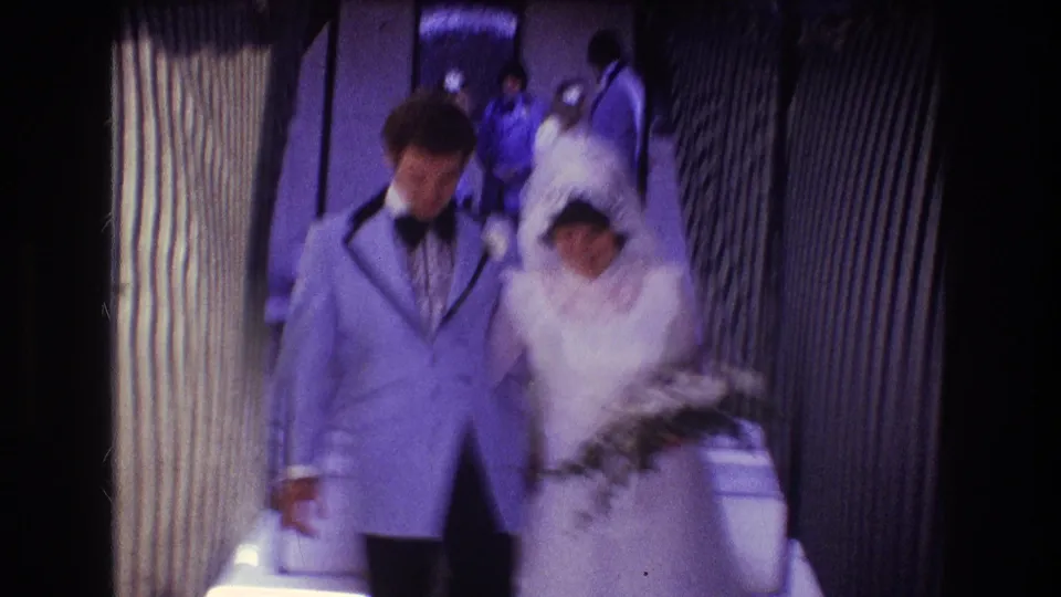 The bride and groom move past the handheld camera in the Boston wedding hallway, with the groom's pale blue jacket and the bride's veil prominent in the frame.