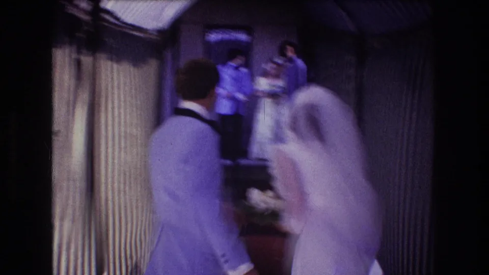 The home-movie camera follows the bride and groom from behind as they turn near the striped corridor, with other wedding guests visible ahead.
