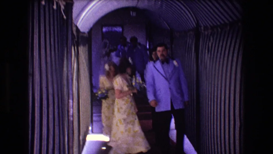 Later in the 1974 Boston wedding hallway scene, formally dressed guests continue through the corridor, including a woman holding flowers and a man in a light blue jacket.