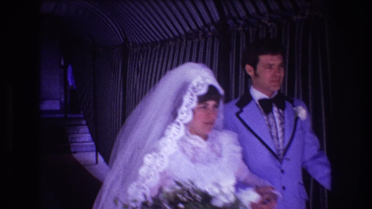 In supported 1974 Boston home-movie footage, a bride in a lace-trimmed veil and a groom in a pale suit walk together through a dim corridor.