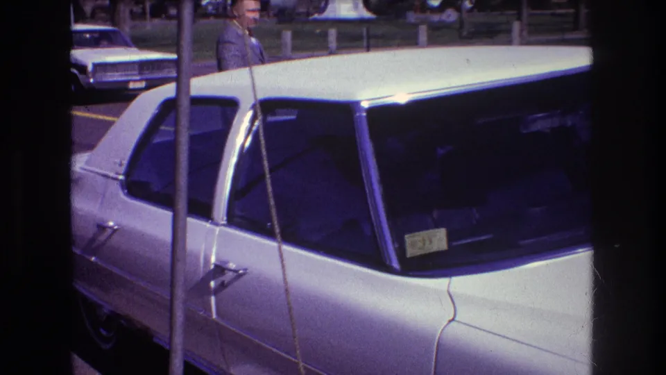 A daytime 1974 Boston home-movie frame shows a white car parked outdoors, with a partially visible person near the vehicle and another car in the background.