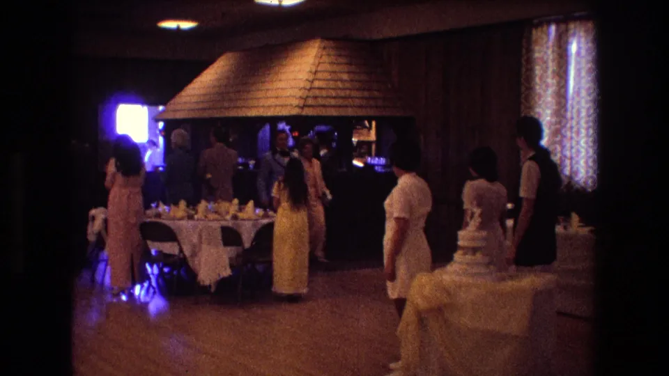 A warmly lit reception room shows guests standing near banquet tables and a tiered wedding cake, with wood-paneled walls and curtains visible.