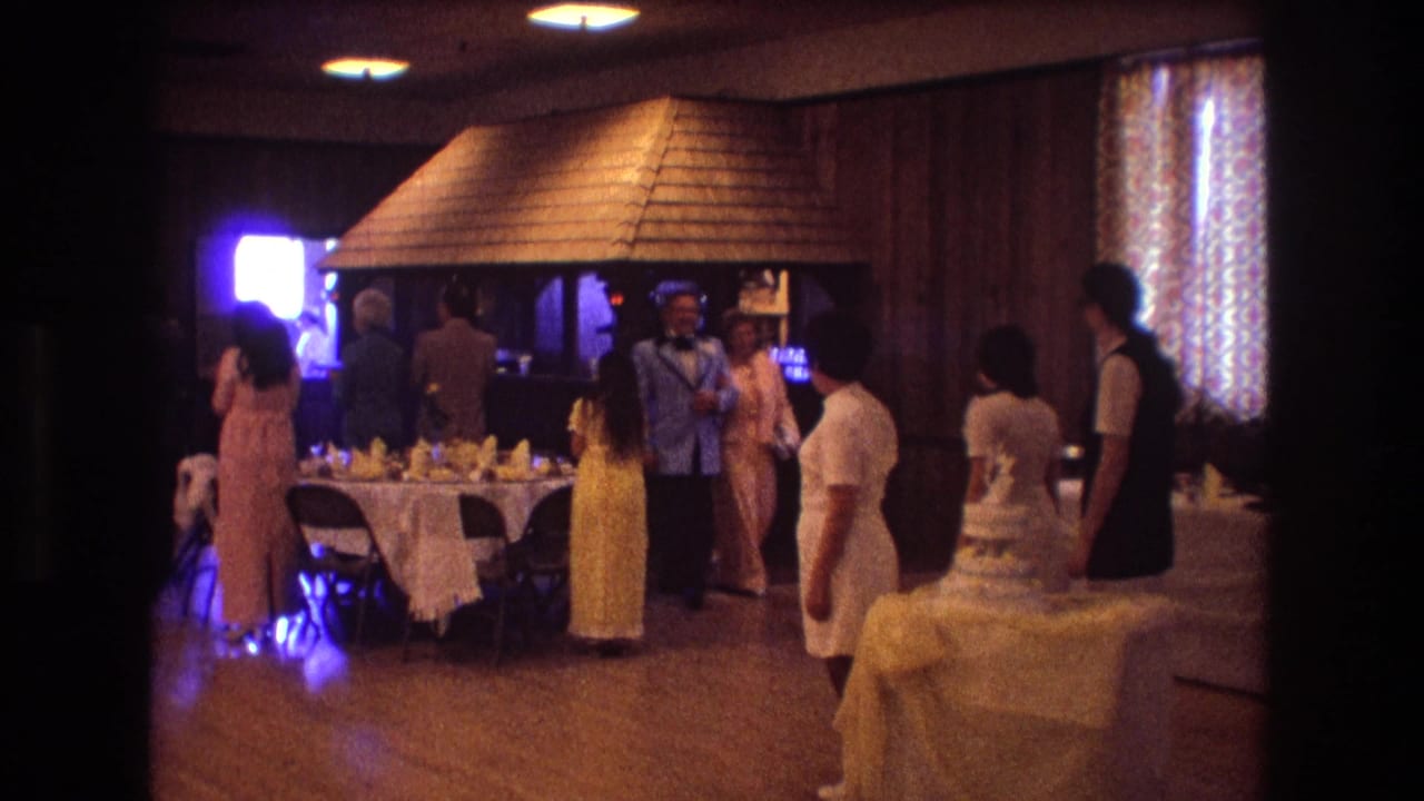 1974 Boston wedding reception guests stand and mingle near a multi-tiered cake table in a warm wood-paneled banquet hall.