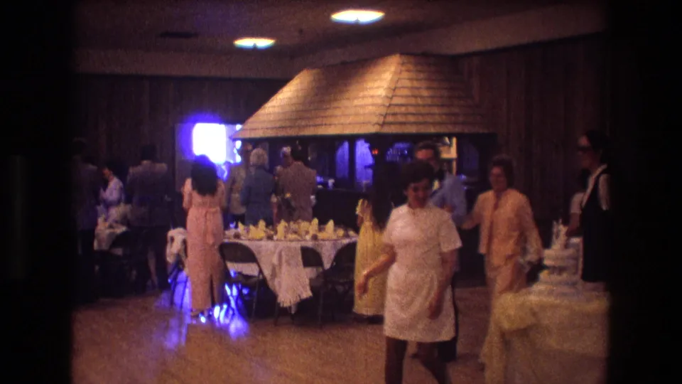 A woman in a light dress walks past the wedding cake during a 1974 Boston reception while guests gather near the banquet tables.