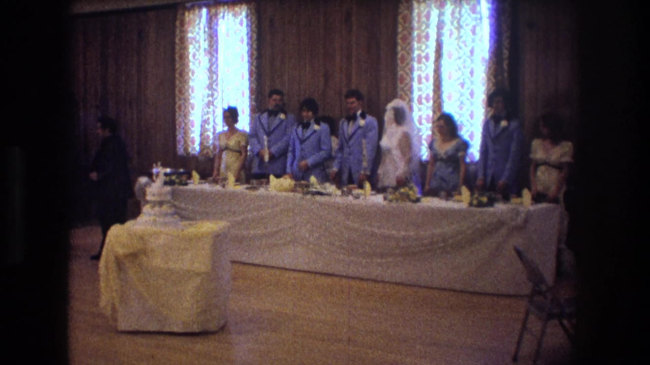 1974 Boston home-movie frame showing a bride, groom, attendants, guests, a tiered cake, and a draped head table inside a wood-paneled wedding reception room.