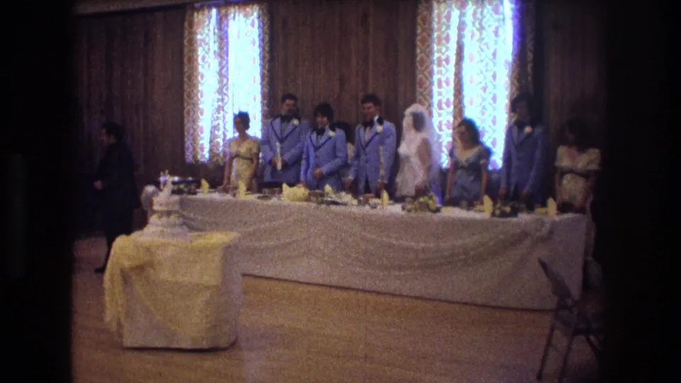 1974 Boston home-movie frame showing a bride, groom, attendants, guests, a tiered cake, and a draped head table inside a wood-paneled wedding reception room.