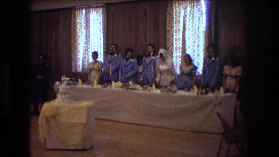 1974 Boston wedding reception frame with the bride and groom among attendants at a long white-draped table, with cake, curtains, and wood-paneled walls visible.