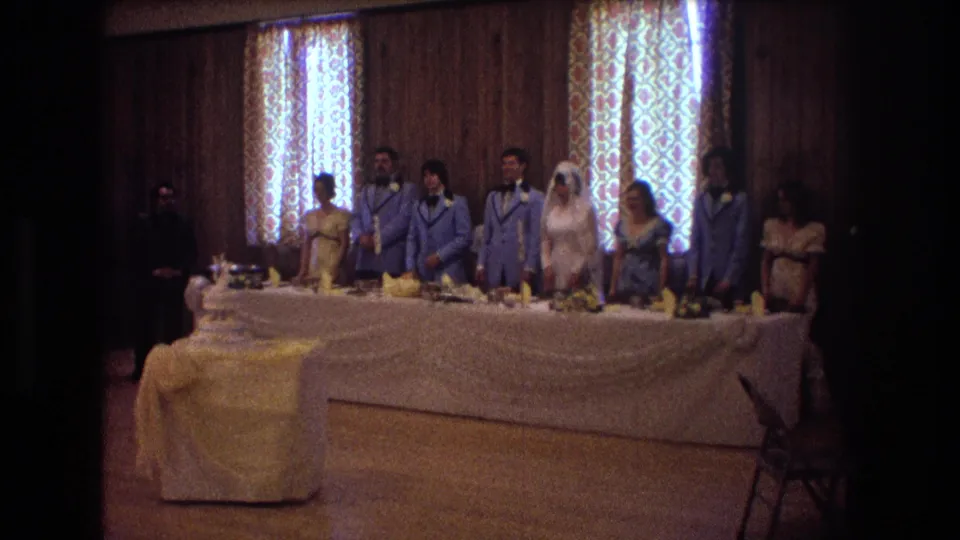 1974 Boston 8mm frame showing the wedding party lined behind the head table, with the bride near center-right and guests at the edges of the reception room.