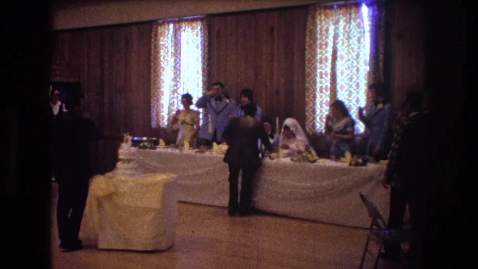 1974 Boston wedding reception frame with a man facing the head table, the bride and groom near the center, attendants and guests around them, and the tiered cake at the table end.