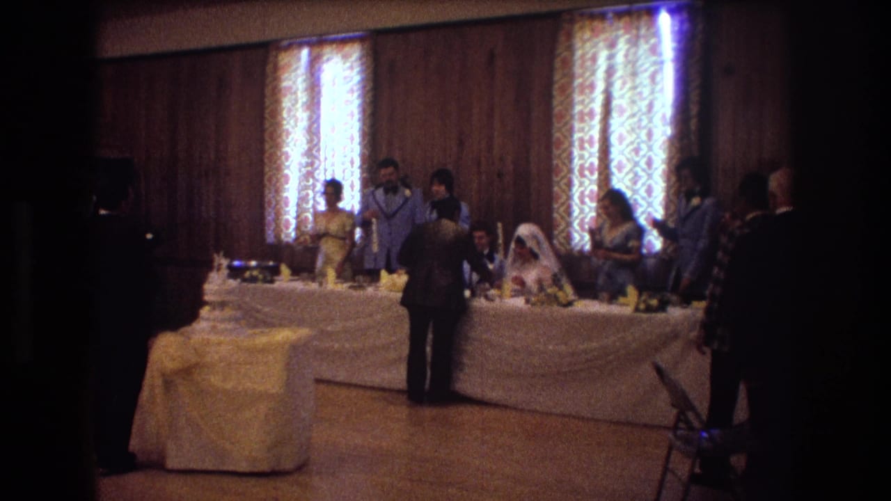 Bride and groom seated at a reception table with guests nearby and a wedding cake on a side table.