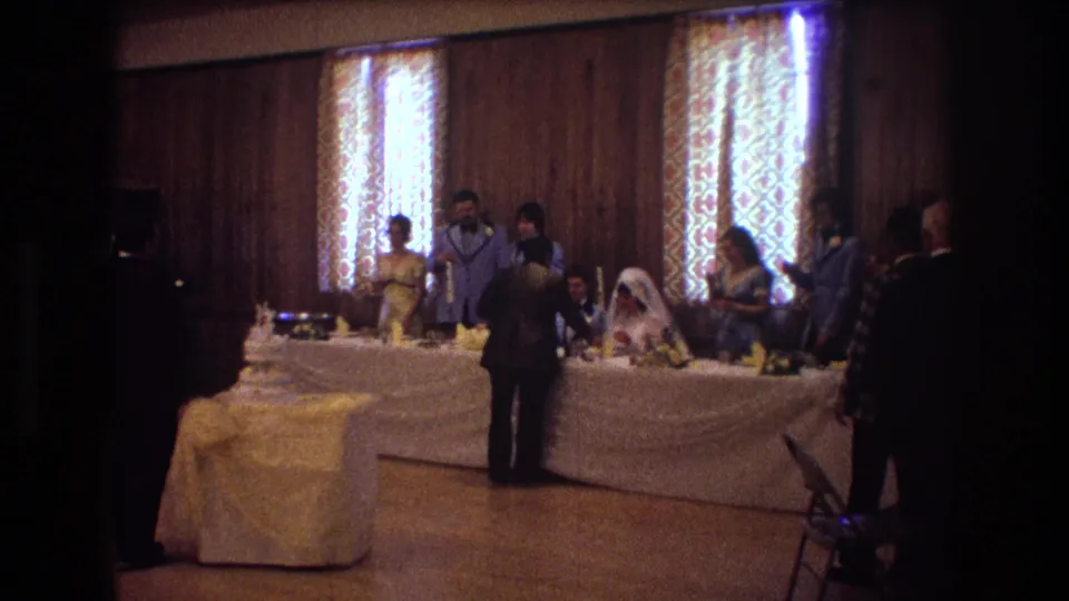 1974 Boston 8mm home-movie frame showing a bride and groom at a decorated wedding reception table as guests stand nearby in a wood-paneled room.