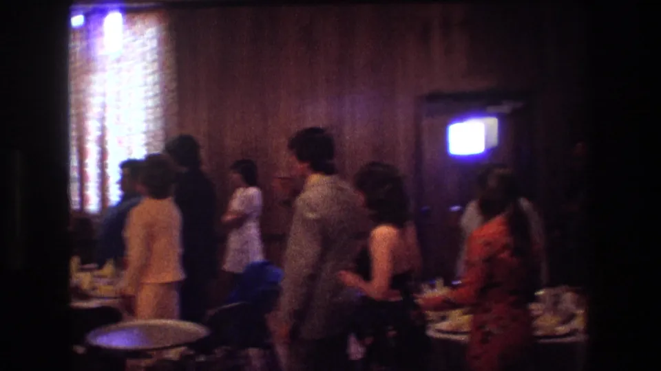 1974 Boston home-movie frame showing reception guests standing along the wall as the camera finishes its pan across the private wedding venue.