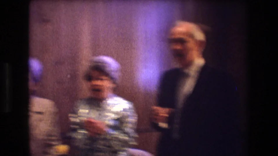 1974 Boston home-movie frame showing a woman in a patterned dress and pillbox-style hat near an older man holding a drink in a dim reception room.