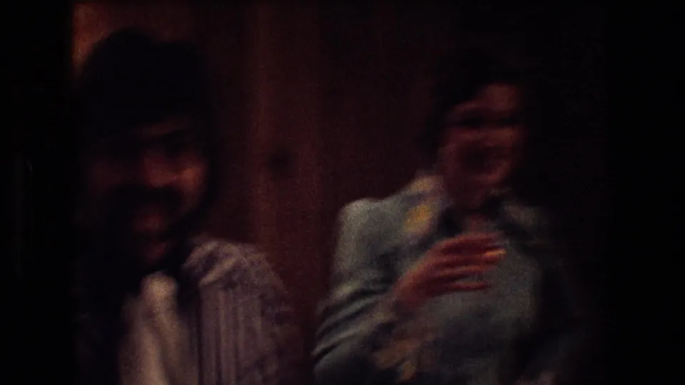 1974 Boston home-movie frame showing two smiling reception guests in low light, including a woman holding a drink, with wood paneling behind them.