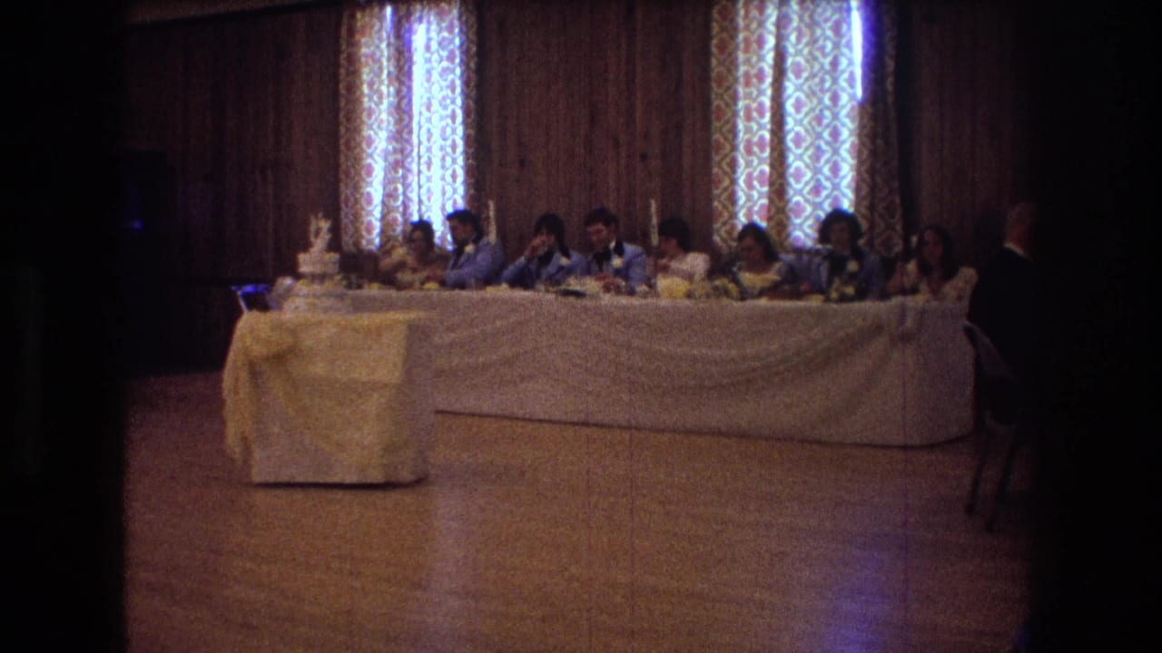1974 Boston home-movie frame showing wedding guests seated along a long white-draped head table in a wood-paneled reception room, with a tiered cake on a covered stand at the left.