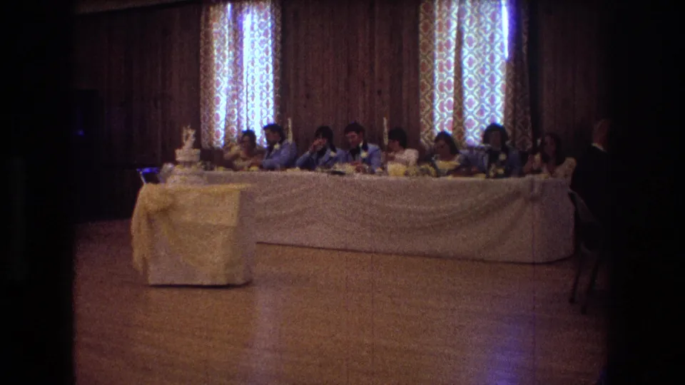 1974 Boston home-movie frame showing wedding guests seated along a long white-draped head table in a wood-paneled reception room, with a tiered cake on a covered stand at the left.