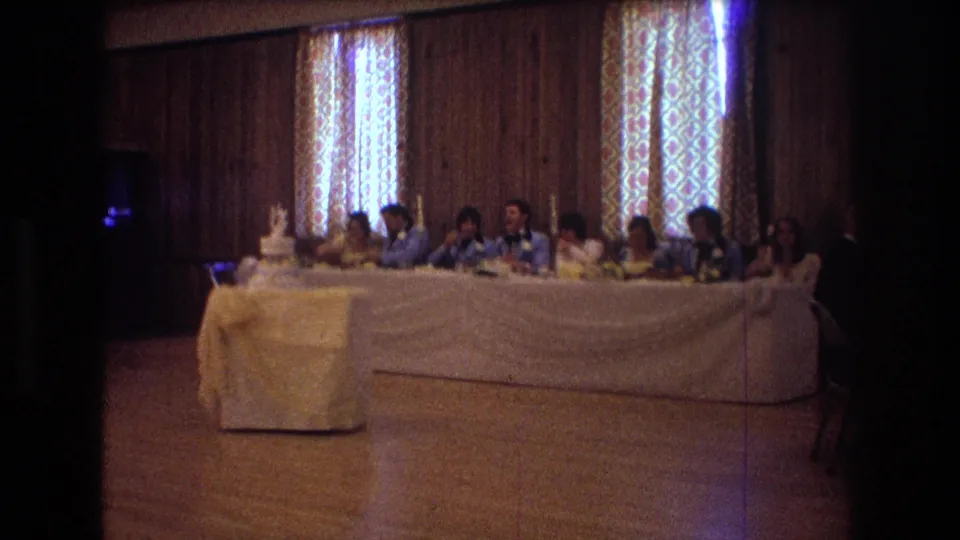 1974 Boston wedding reception frame with guests seated at a white-draped head table, the wedding cake at the left, and patterned curtains filtering light behind them.