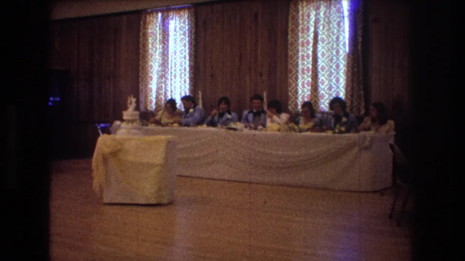 1974 Boston home-movie frame showing a stationary view of wedding guests at a long head table, with a tiered cake on the left and tall patterned curtains behind the group.