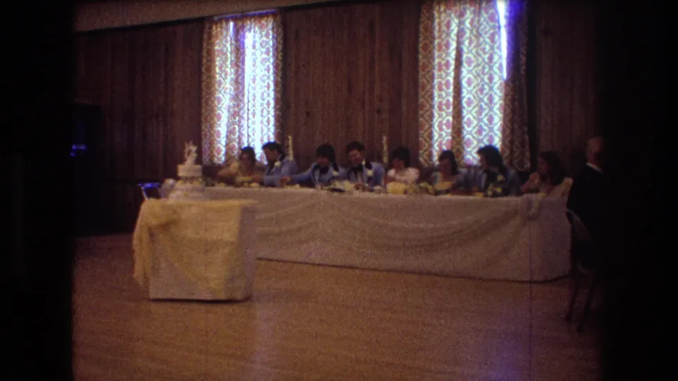 1974 Boston wedding reception frame showing guests seated along the head table, the cake at the left, and a standing figure near the right edge inside the wood-paneled hall.