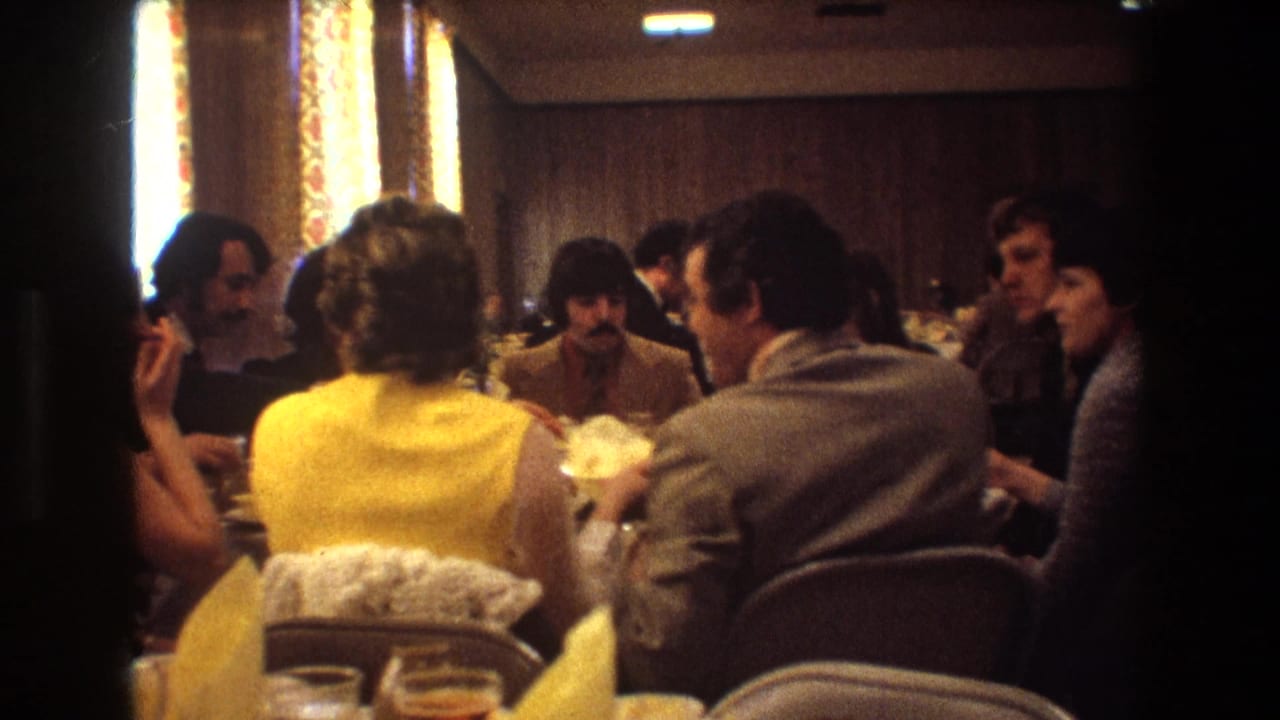 Guests at a 1974 Boston wedding reception sit closely around a table with plates and glasses in a warm, wood-paneled room.