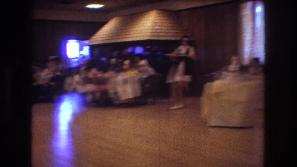 A soft handheld frame shows a large reception room with guests seated along the side and a woman in a white dress moving through the open floor.