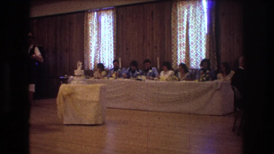 The head table at the 1974 Boston wedding reception is shown with seated guests, floral arrangements, a wedding cake, and patterned curtains behind them.