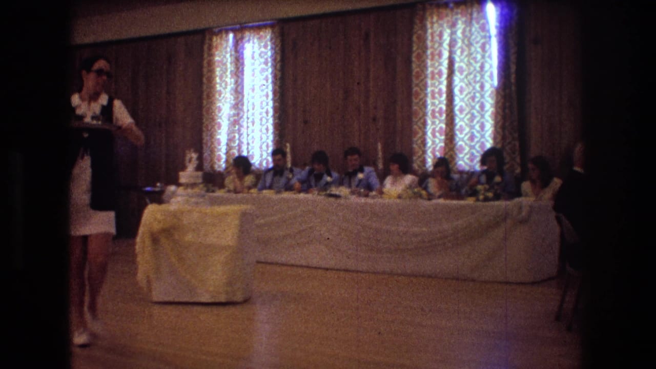 1974 Boston home-movie frame showing a waitress carrying a tray past a long reception table, with guests seated behind white linens and a tiered wedding cake on a yellow-covered stand.