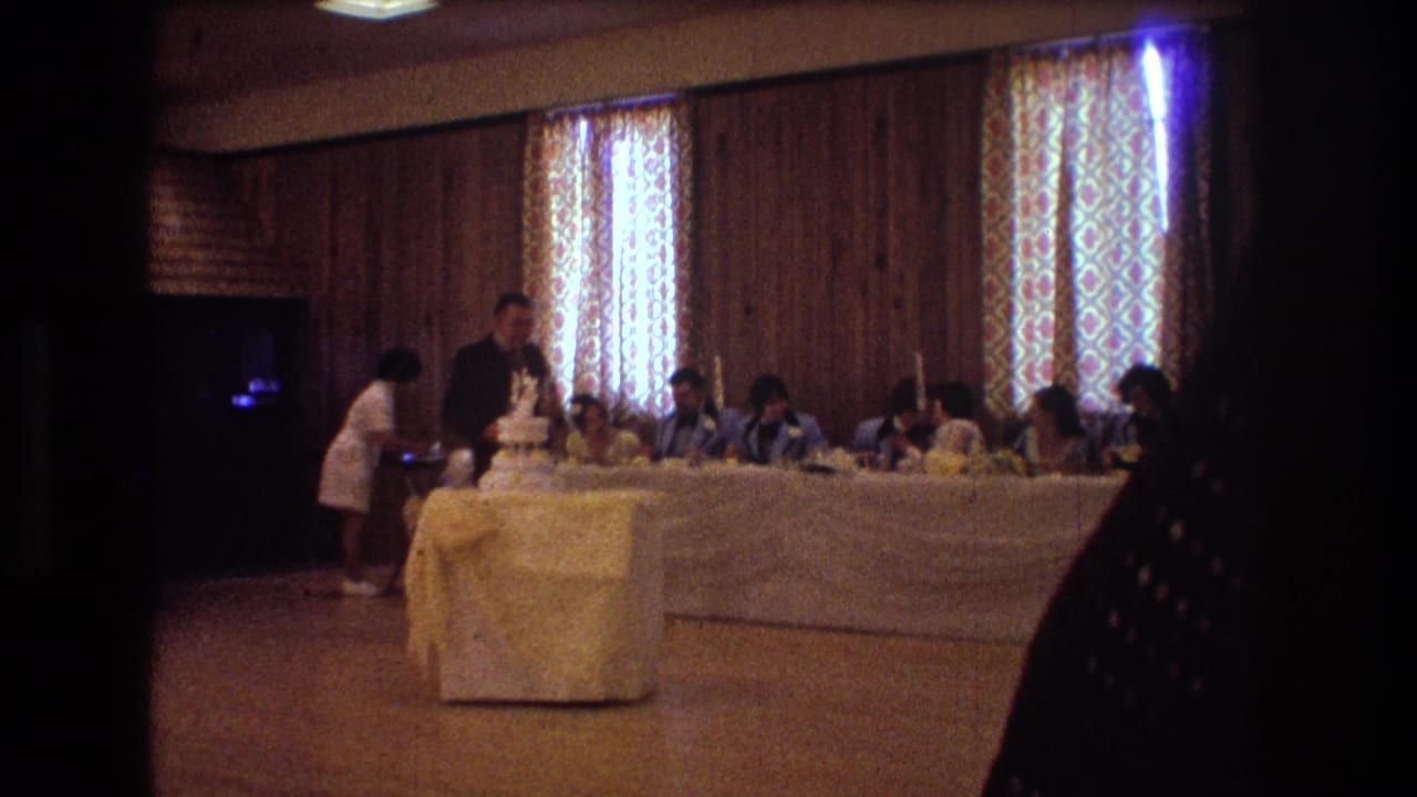 Suited man stands near a tiered wedding cake while guests sit at a long reception table.