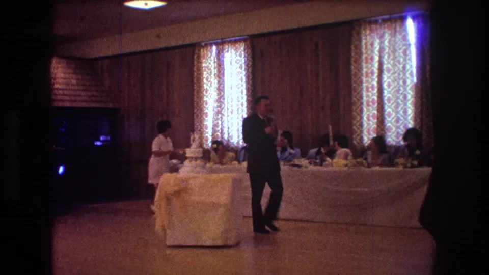 1974 Boston home-movie frame showing a man in a dark suit crossing the reception floor in front of a tiered wedding cake, with guests seated at a long table in the background.