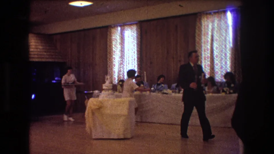 1974 Boston reception frame with a suited man walking across the foreground, a waitress near the cake table, and seated guests visible against patterned curtains and wood-paneled walls.