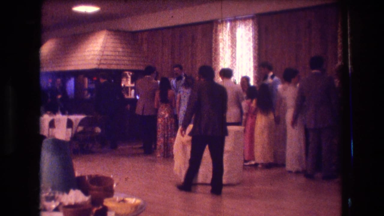 1974 Boston home-movie frame showing guests in suits and dresses standing near a function-room floor with wood paneling and patterned curtains.