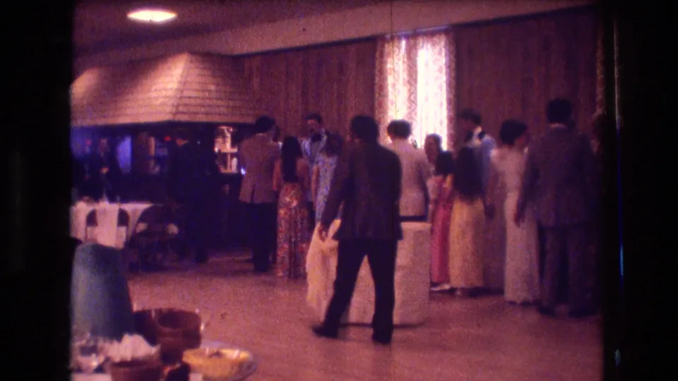1974 Boston home-movie frame showing guests in suits and dresses standing near a function-room floor with wood paneling and patterned curtains.