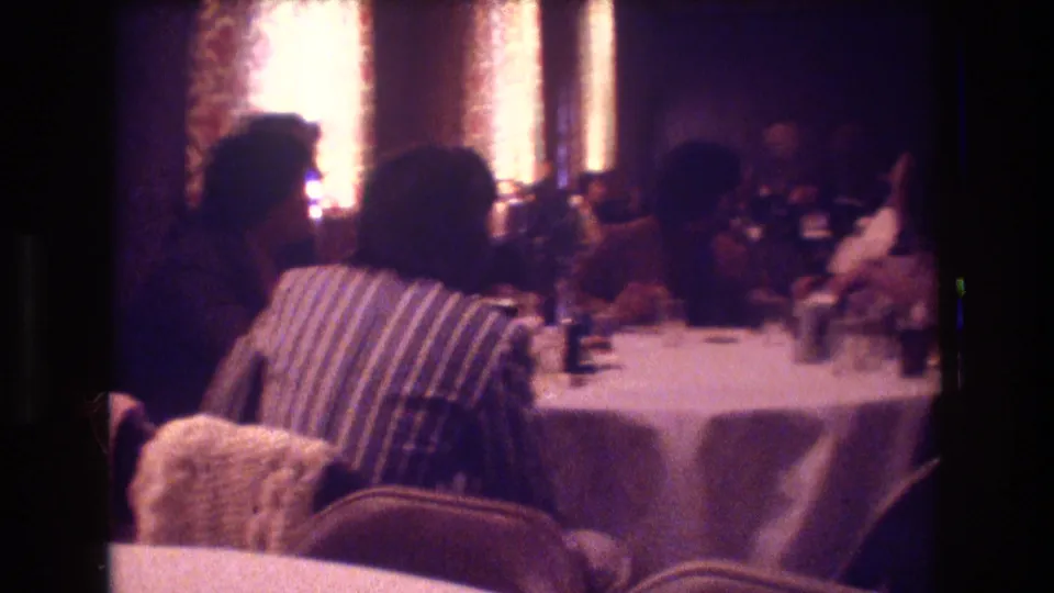 1974 Boston home-movie frame from table level showing seated guests around a white-clothed banquet table in a dim indoor reception room.