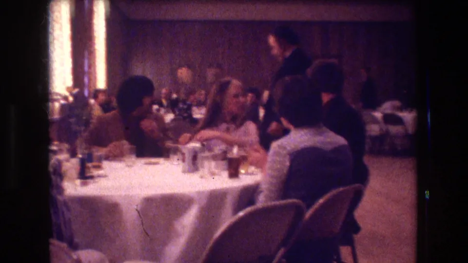 1974 Boston home-movie frame showing people seated at a banquet table with drinks and tableware as a standing figure appears behind them in the function room.