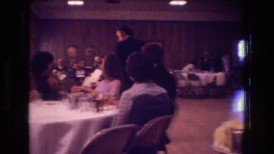 1974 Boston home-movie frame showing seated reception guests at a round table with a standing man in the background and wood-paneled walls behind them.