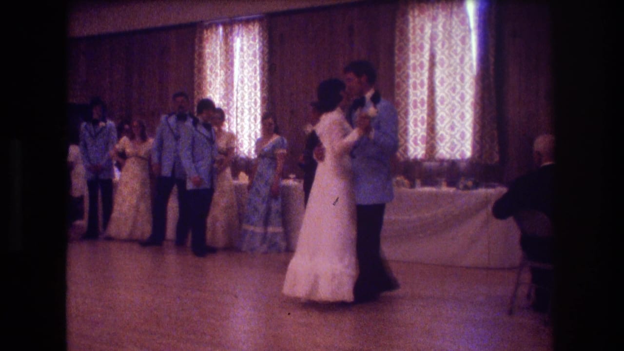 Bride and groom slow dance in a reception hall while attendants and guests stand nearby.