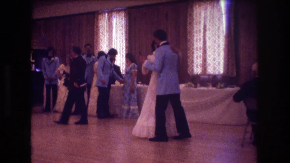 From a handheld guest viewpoint, the groom's light blue jacket and the bride's white gown are seen as they dance near attendants and guests in a warmly lit private reception room.