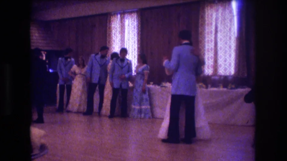 Near the end of the clip, the groom in a light blue jacket stands in the foreground while attendants and guests cluster by the covered table under patterned curtains in the reception hall.