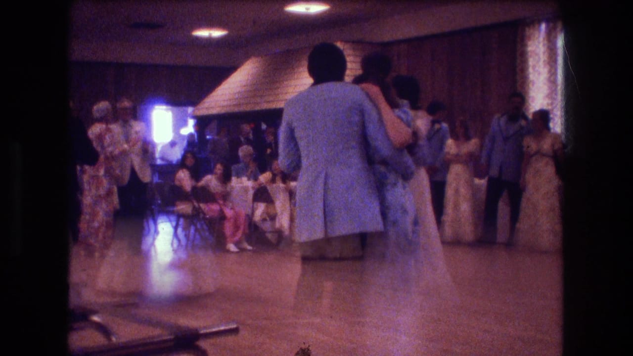 In 1974 Boston home-movie footage, wedding guests in pale dresses and light blue jackets waltz on a wooden dance floor inside a wood-paneled reception room.