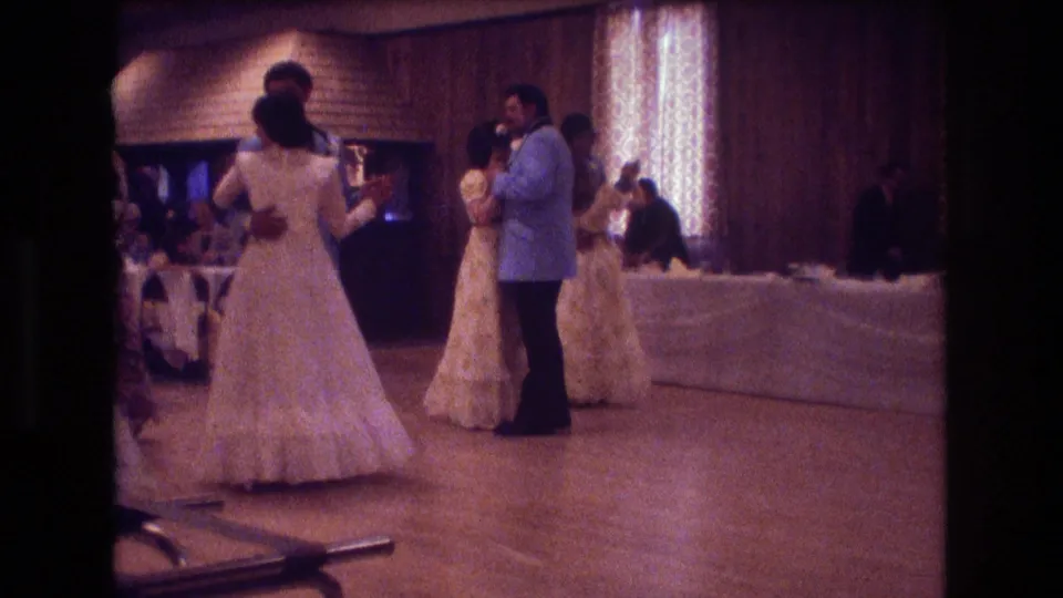 A couple in formal wedding reception attire waltzes near other dancers and a white-draped table in the Boston 1974 home-movie scene.