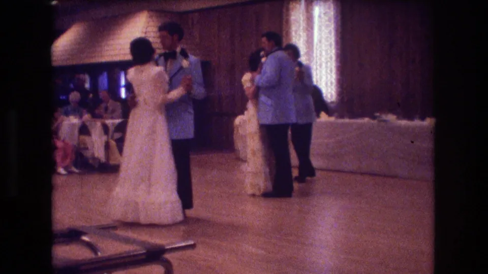 A foreground couple waltzes on the wooden floor as additional wedding guests stand and sit in the softly lit 1974 Boston reception interior.