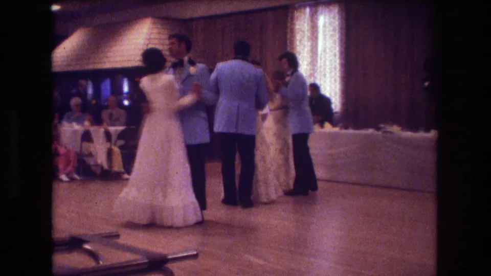 Several formally dressed couples dance together beside a long white-covered reception table in the warm, grainy 1974 Boston wedding footage.