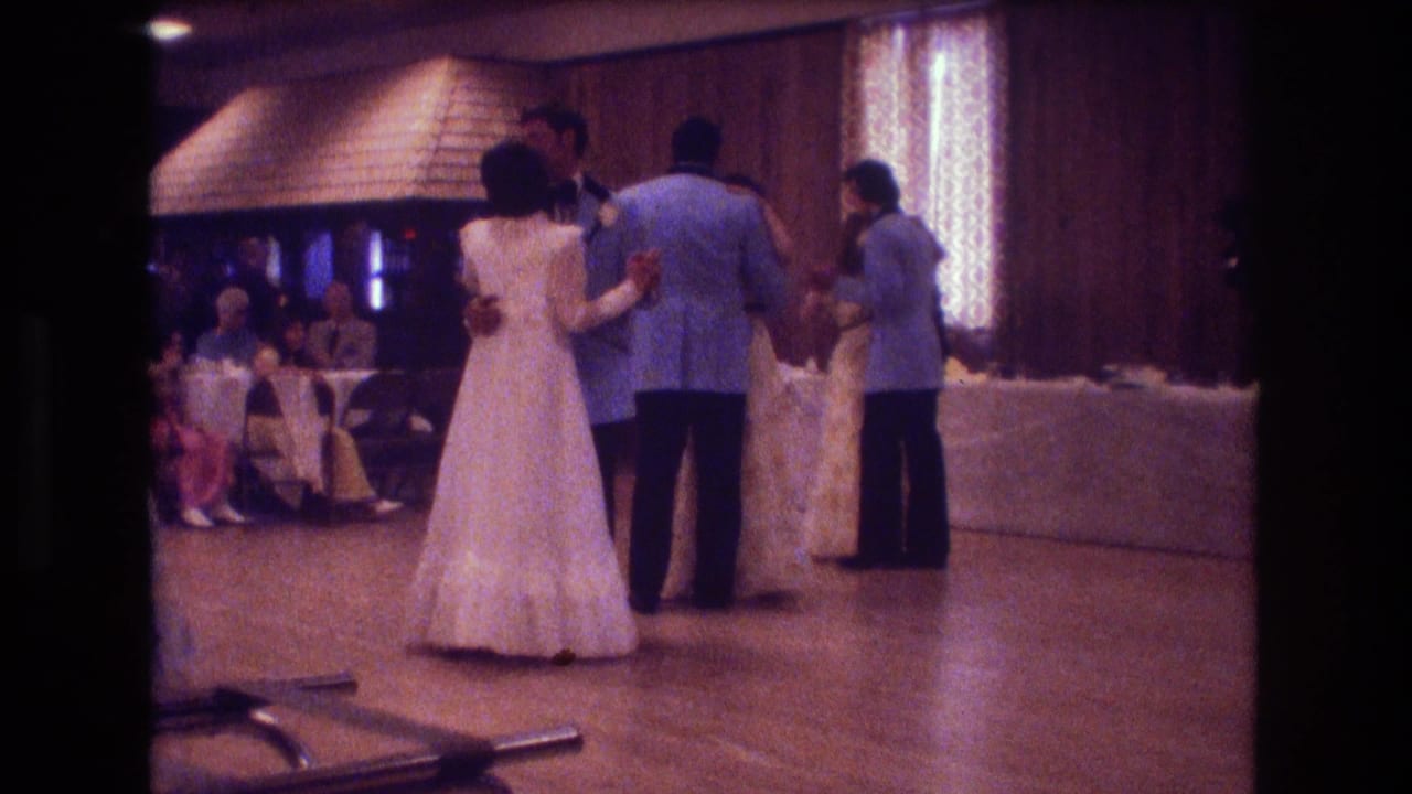 In this 1974 Boston home-movie frame, formally dressed couples dance at a wedding reception under warm lights with wood-paneled walls and seated guests behind them.