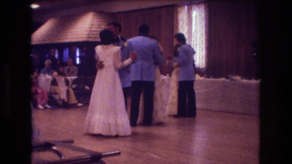 In this 1974 Boston home-movie frame, formally dressed couples dance at a wedding reception under warm lights with wood-paneled walls and seated guests behind them.