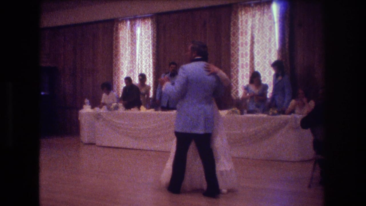 Bride and groom dancing on a wooden floor with guests watching from banquet tables.